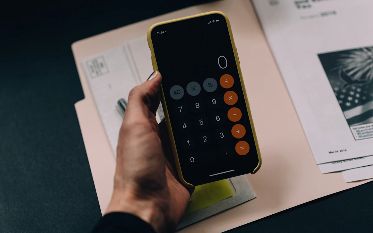 Una mano sostiene un celular con la calculadora abierta sobre una carpeta con documentos