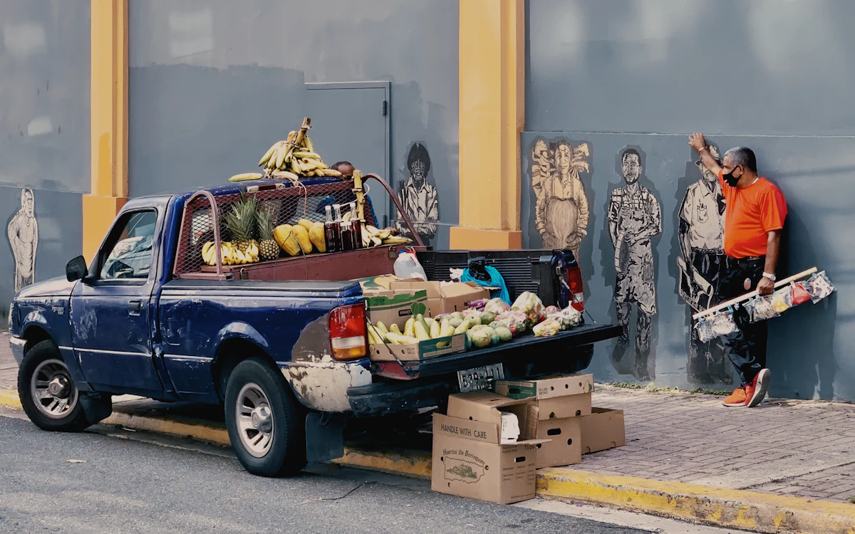 Pick-up azul con la caja cargada de fruta, estacionada en una banqueta junto a un vendedor